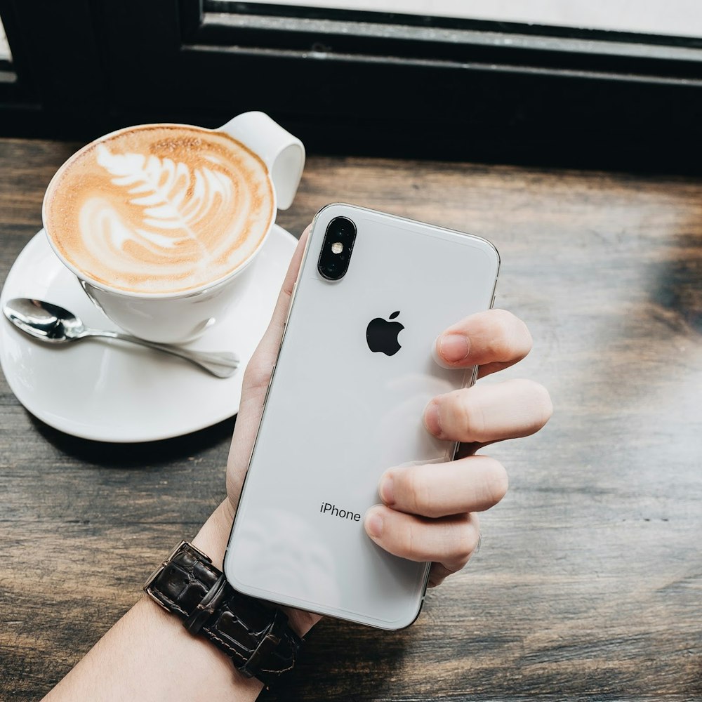 person holding gold iPhone Xs near cappuccino beside gray stainless steel teaspoon