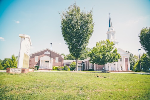A church with a tall steeple sits among trees and greenery under a clear blue sky. The building has a white facade with brick accents, and a large sign is visible on the grass in the foreground.