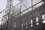 A black and white image of a construction site featuring a facade of a classical building being supported by scaffolding. The intricate architectural details of the building are visible through a translucent mesh, providing a contrast between the old-style architecture and the modern construction framework.