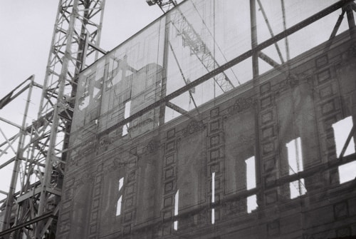 A black and white image of a construction site featuring a facade of a classical building being supported by scaffolding. The intricate architectural details of the building are visible through a translucent mesh, providing a contrast between the old-style architecture and the modern construction framework.