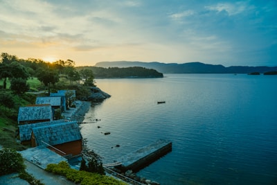 Several small, rustic cottages with blue-tiled roofs line the shore of a calm, expansive lake. The sun is setting behind distant mountains, casting a warm glow over the landscape. Trees and bushes are scattered along the rocky shoreline, and a lone boat floats in the water, creating a serene and picturesque scene.