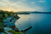 Several small, rustic cottages with blue-tiled roofs line the shore of a calm, expansive lake. The sun is setting behind distant mountains, casting a warm glow over the landscape. Trees and bushes are scattered along the rocky shoreline, and a lone boat floats in the water, creating a serene and picturesque scene.