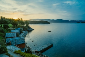 Several small, rustic cottages with blue-tiled roofs line the shore of a calm, expansive lake. The sun is setting behind distant mountains, casting a warm glow over the landscape. Trees and bushes are scattered along the rocky shoreline, and a lone boat floats in the water, creating a serene and picturesque scene.