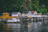 A row of charming waterside cabins stands on wooden stilts over a calm body of water. The cabins vary in design, with some painted white and others in shades of yellow. A lush green forest serves as a backdrop, adding to the serene atmosphere. Bright orange buoys float on the water's surface, and a stone wall supports the boardwalk leading to the cabins.