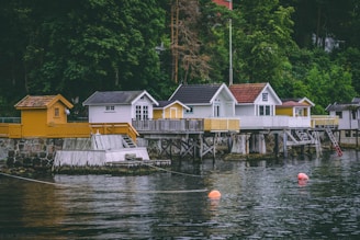 A row of charming waterside cabins stands on wooden stilts over a calm body of water. The cabins vary in design, with some painted white and others in shades of yellow. A lush green forest serves as a backdrop, adding to the serene atmosphere. Bright orange buoys float on the water's surface, and a stone wall supports the boardwalk leading to the cabins.