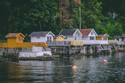 A row of charming waterside cabins stands on wooden stilts over a calm body of water. The cabins vary in design, with some painted white and others in shades of yellow. A lush green forest serves as a backdrop, adding to the serene atmosphere. Bright orange buoys float on the water's surface, and a stone wall supports the boardwalk leading to the cabins.