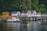 A row of charming waterside cabins stands on wooden stilts over a calm body of water. The cabins vary in design, with some painted white and others in shades of yellow. A lush green forest serves as a backdrop, adding to the serene atmosphere. Bright orange buoys float on the water's surface, and a stone wall supports the boardwalk leading to the cabins.