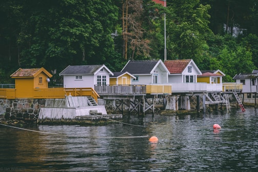 A row of charming waterside cabins stands on wooden stilts over a calm body of water. The cabins vary in design, with some painted white and others in shades of yellow. A lush green forest serves as a backdrop, adding to the serene atmosphere. Bright orange buoys float on the water's surface, and a stone wall supports the boardwalk leading to the cabins.