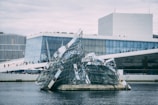 A modern architectural building with angular, glass-filled designs rising from the water in front. The main structure has clean geometric lines and large glass windows. People can be seen walking along the sloped roof area, and the sky appears overcast.