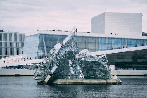 A modern architectural building with angular, glass-filled designs rising from the water in front. The main structure has clean geometric lines and large glass windows. People can be seen walking along the sloped roof area, and the sky appears overcast.