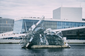 A modern architectural building with angular, glass-filled designs rising from the water in front. The main structure has clean geometric lines and large glass windows. People can be seen walking along the sloped roof area, and the sky appears overcast.