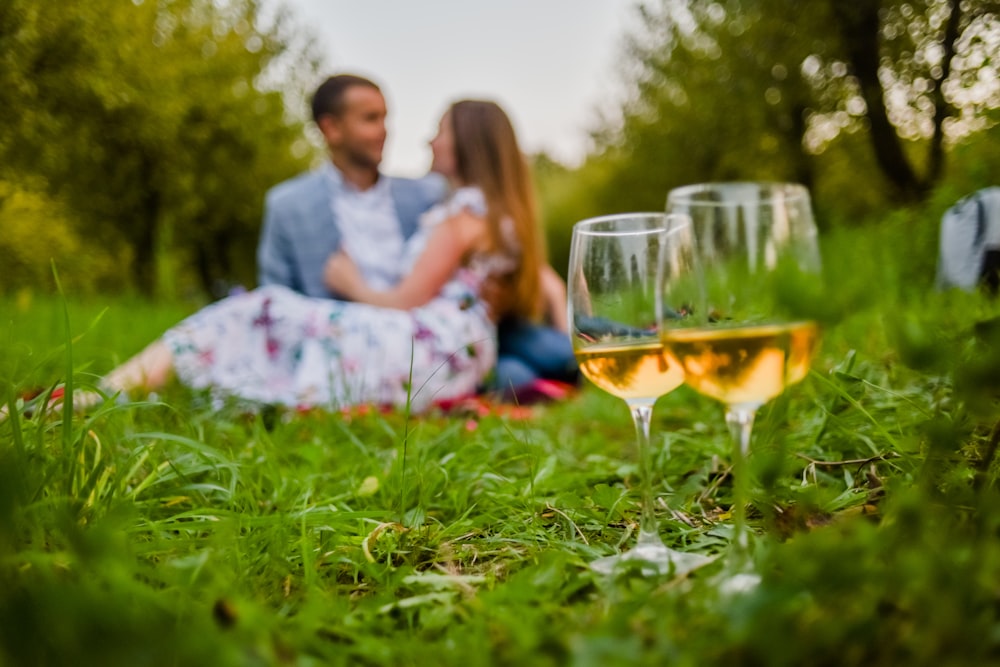 a man and a woman sitting in the grass with a glass of wine