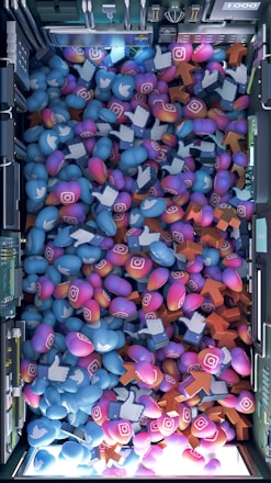 A top-down view of a vibrant pile of colorful icons and symbols representing various social media platforms. Icons include blue Twitter birds, Instagram logos, Facebook-like thumbs up, and orange up arrows. The symbols are situated inside a futuristic, mechanical enclosure, possibly resembling a server or machine.