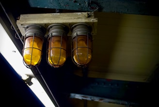 Industrial-style light fixtures with protective cages enclosing orange-tinted bulbs are mounted on a weathered ceiling. The surrounding area is dimly lit, adding to the utilitarian and somewhat aged atmosphere.