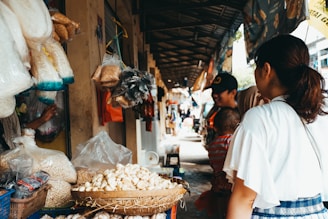 A bustling outdoor market scene where a variety of goods are displayed, including plastic bags of grains, baskets of garlic, and other groceries. A woman and child are seen interacting with a vendor, while other people are walking along the covered aisle.