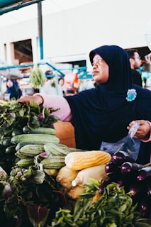 A person wearing a dark headscarf is selecting fresh vegetables at a market. Various vegetables, including corn, cucumbers, and eggplants, are displayed on the stall. The setting appears vibrant and busy, with other people and market items visible in the background.