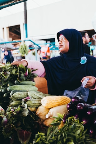 A person wearing a dark headscarf is selecting fresh vegetables at a market. Various vegetables, including corn, cucumbers, and eggplants, are displayed on the stall. The setting appears vibrant and busy, with other people and market items visible in the background.