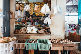 A bustling market stall displays a colorful array of fresh produce and packaged goods. Various vegetables and spices are arranged in baskets and crates, with plastic-wrapped items hanging above. The stall is rich with earthy textures and vibrant colors, reflecting a lively market atmosphere. A vendor is partly visible in the background, attending to the arrangement.