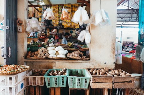 A bustling market stall displays a colorful array of fresh produce and packaged goods. Various vegetables and spices are arranged in baskets and crates, with plastic-wrapped items hanging above. The stall is rich with earthy textures and vibrant colors, reflecting a lively market atmosphere. A vendor is partly visible in the background, attending to the arrangement.