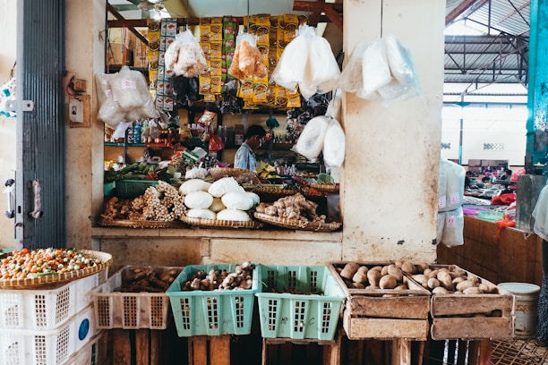 A bustling market stall displays a colorful array of fresh produce and packaged goods. Various vegetables and spices are arranged in baskets and crates, with plastic-wrapped items hanging above. The stall is rich with earthy textures and vibrant colors, reflecting a lively market atmosphere. A vendor is partly visible in the background, attending to the arrangement.