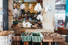 A bustling market stall displays a colorful array of fresh produce and packaged goods. Various vegetables and spices are arranged in baskets and crates, with plastic-wrapped items hanging above. The stall is rich with earthy textures and vibrant colors, reflecting a lively market atmosphere. A vendor is partly visible in the background, attending to the arrangement.