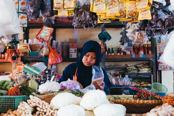 A woman wearing a dark blue headscarf is focused on preparing or packaging goods in a small shop filled with various food items. The shelves behind her are stocked with packaged goods such as spices, sauces, and snacks. Fresh produce, including leafy greens and root vegetables, is displayed in baskets in front of her.