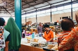 A bustling indoor market with various stalls displaying fresh produce and goods. Several people are visible interacting with vendors or browsing items. The market is housed in a large, open warehouse with a high ceiling and metal beams.