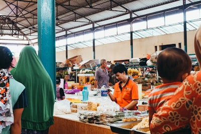 A bustling indoor market with various stalls displaying fresh produce and goods. Several people are visible interacting with vendors or browsing items. The market is housed in a large, open warehouse with a high ceiling and metal beams.