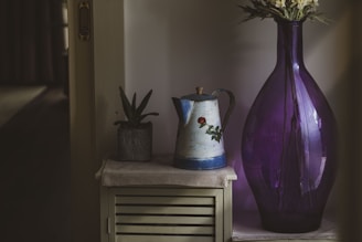 A large, translucent purple vase with flowers is placed next to a vintage-style metal pitcher with floral design and a small potted plant. They are arranged on a wooden cabinet with a white cloth, creating a cozy, rustic atmosphere.