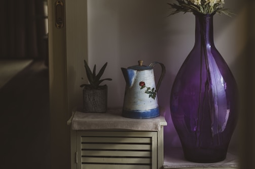A large, translucent purple vase with flowers is placed next to a vintage-style metal pitcher with floral design and a small potted plant. They are arranged on a wooden cabinet with a white cloth, creating a cozy, rustic atmosphere.