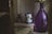 A large, translucent purple vase with flowers is placed next to a vintage-style metal pitcher with floral design and a small potted plant. They are arranged on a wooden cabinet with a white cloth, creating a cozy, rustic atmosphere.