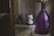 A large, translucent purple vase with flowers is placed next to a vintage-style metal pitcher with floral design and a small potted plant. They are arranged on a wooden cabinet with a white cloth, creating a cozy, rustic atmosphere.
