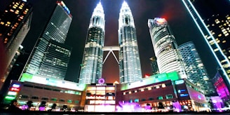 A night scene featuring the brightly lit Petronas Twin Towers in the center surrounded by modern skyscrapers. The lower part of the image shows a building with vibrant neon lights. The sky is dark, providing a stark contrast to the illuminated structures.