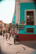 A vibrant street scene in a city with colorful buildings, featuring turquoise and red walls. Several people are walking along the street, and there are signs indicating directions to various locations like Meson del Rosario and Valenciana. The atmosphere suggests a lively and bustling urban setting.