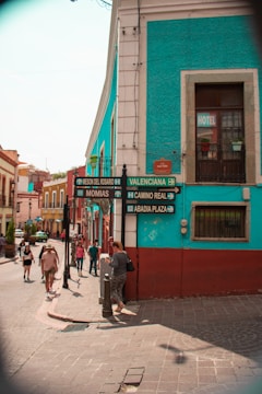 A vibrant street scene in a city with colorful buildings, featuring turquoise and red walls. Several people are walking along the street, and there are signs indicating directions to various locations like Meson del Rosario and Valenciana. The atmosphere suggests a lively and bustling urban setting.
