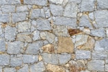 An arrangement of roughly cut gray and brown stones fitted together to form a wall. The stones vary in size and shape, with some having smoother edges while others have a more jagged appearance. The mortar used to hold the stones together is visible in the gaps between them.