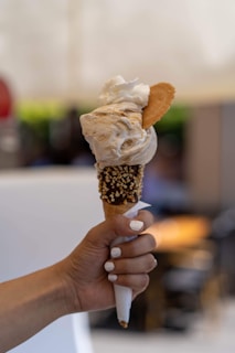 A hand with white nail polish holds an ice cream cone topped with a large scoop of creamy ice cream, a dollop of whipped cream, and a small biscuit. The cone is decorated with a chocolate coating and sprinkled with chopped nuts. The background is blurred, suggesting an indoor or outdoor setting with soft lighting.