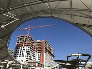 A construction site with a partially completed high-rise building. Two cranes are actively working on the site. The scene is framed by a large, curved structure that appears to be part of a covered walkway or roof. The sky is clear with a deep blue color.