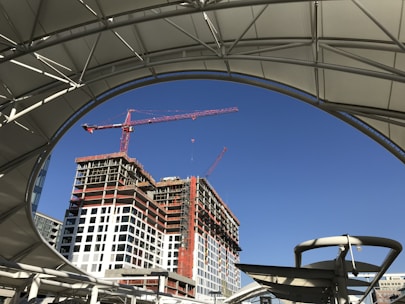 A construction site with a partially completed high-rise building. Two cranes are actively working on the site. The scene is framed by a large, curved structure that appears to be part of a covered walkway or roof. The sky is clear with a deep blue color.