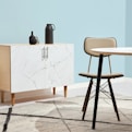 A minimalist interior space features a light wood cabinet with a white marble pattern on its doors, adorned with two matte black decorative vases. A modern chair with a tan cushion sits next to a round table with a similar light marble top. The setting is complemented by a pale blue wall and a geometric-patterned rug that incorporates shades of grey and cream.