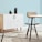 A minimalist interior space features a light wood cabinet with a white marble pattern on its doors, adorned with two matte black decorative vases. A modern chair with a tan cushion sits next to a round table with a similar light marble top. The setting is complemented by a pale blue wall and a geometric-patterned rug that incorporates shades of grey and cream.