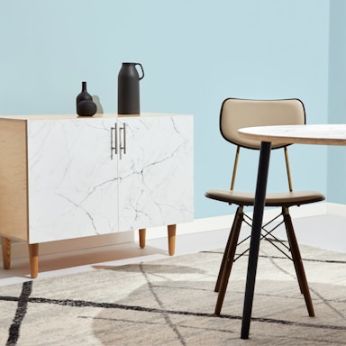 A minimalist interior space features a light wood cabinet with a white marble pattern on its doors, adorned with two matte black decorative vases. A modern chair with a tan cushion sits next to a round table with a similar light marble top. The setting is complemented by a pale blue wall and a geometric-patterned rug that incorporates shades of grey and cream.