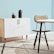 A minimalist interior space features a light wood cabinet with a white marble pattern on its doors, adorned with two matte black decorative vases. A modern chair with a tan cushion sits next to a round table with a similar light marble top. The setting is complemented by a pale blue wall and a geometric-patterned rug that incorporates shades of grey and cream.