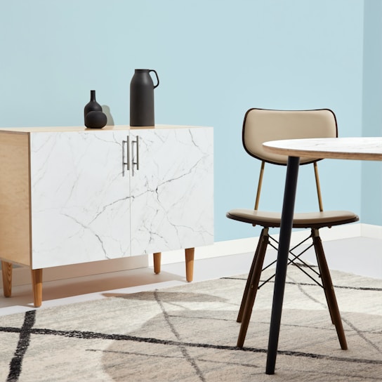 A minimalist interior space features a light wood cabinet with a white marble pattern on its doors, adorned with two matte black decorative vases. A modern chair with a tan cushion sits next to a round table with a similar light marble top. The setting is complemented by a pale blue wall and a geometric-patterned rug that incorporates shades of grey and cream.
