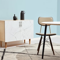 A minimalist interior space features a light wood cabinet with a white marble pattern on its doors, adorned with two matte black decorative vases. A modern chair with a tan cushion sits next to a round table with a similar light marble top. The setting is complemented by a pale blue wall and a geometric-patterned rug that incorporates shades of grey and cream.
