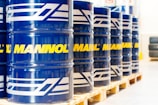 A row of large blue barrels is neatly aligned on wooden pallets. Each barrel prominently features the word 'MANNOL' in bright yellow, with additional white graphics. The scene suggests an industrial or warehouse setting with stacked tires visible in the background.