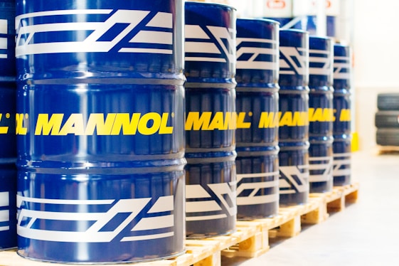 A row of large blue barrels is neatly aligned on wooden pallets. Each barrel prominently features the word 'MANNOL' in bright yellow, with additional white graphics. The scene suggests an industrial or warehouse setting with stacked tires visible in the background.