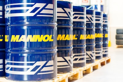 A row of large blue barrels is neatly aligned on wooden pallets. Each barrel prominently features the word 'MANNOL' in bright yellow, with additional white graphics. The scene suggests an industrial or warehouse setting with stacked tires visible in the background.
