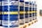 A row of large blue barrels is neatly aligned on wooden pallets. Each barrel prominently features the word 'MANNOL' in bright yellow, with additional white graphics. The scene suggests an industrial or warehouse setting with stacked tires visible in the background.
