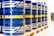 A row of large blue barrels is neatly aligned on wooden pallets. Each barrel prominently features the word 'MANNOL' in bright yellow, with additional white graphics. The scene suggests an industrial or warehouse setting with stacked tires visible in the background.